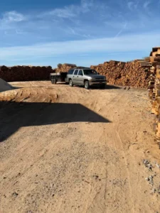 Pickup and dump trailer beside long stacked rows of firewood at the wood yard
