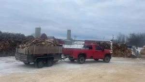 Red pickup and dump trailer loaded with firewood on a snowy yard beside stacked log rows